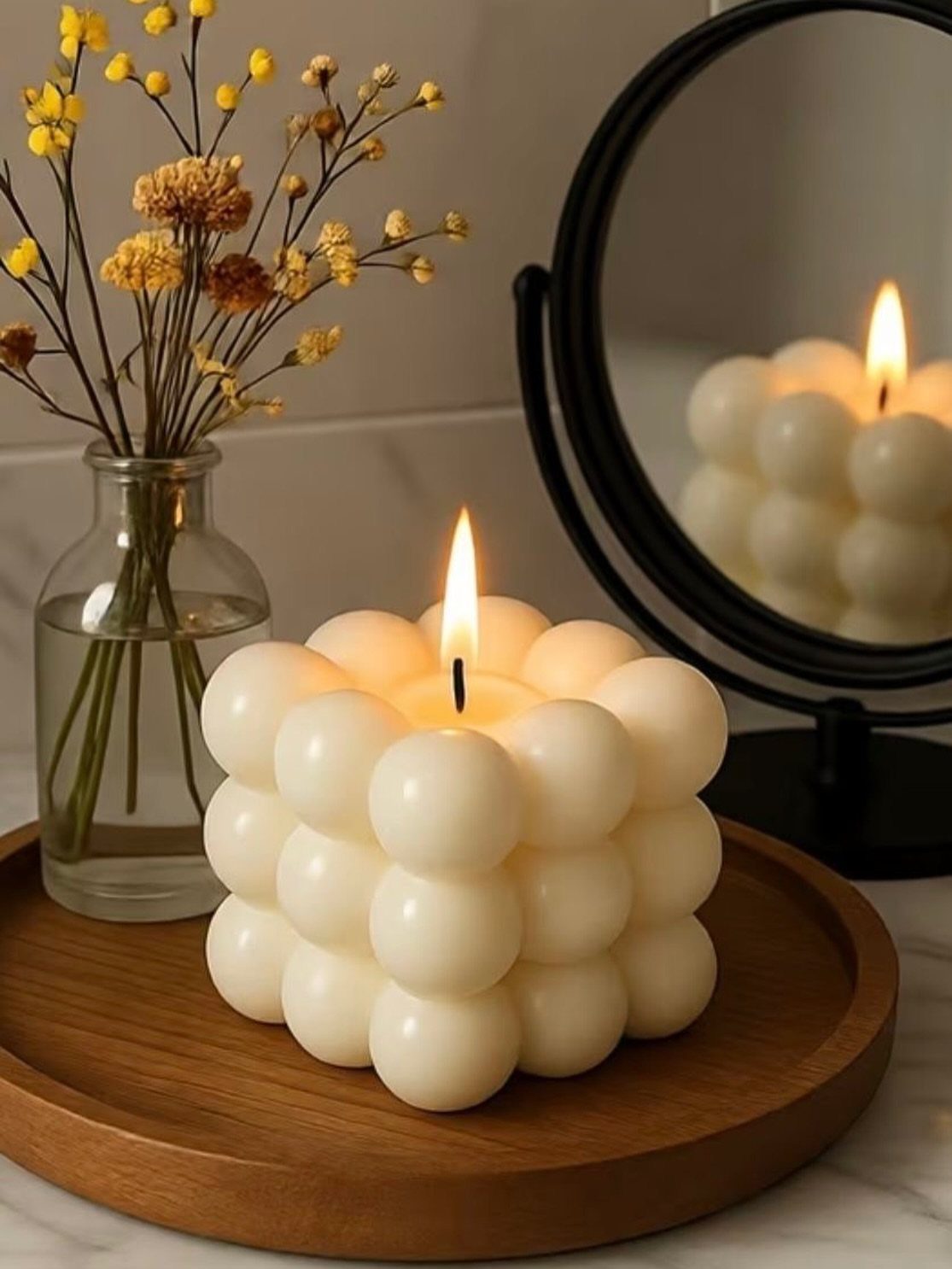 White bubble candle lit on a wooden tray next to yellow dried flowers.
