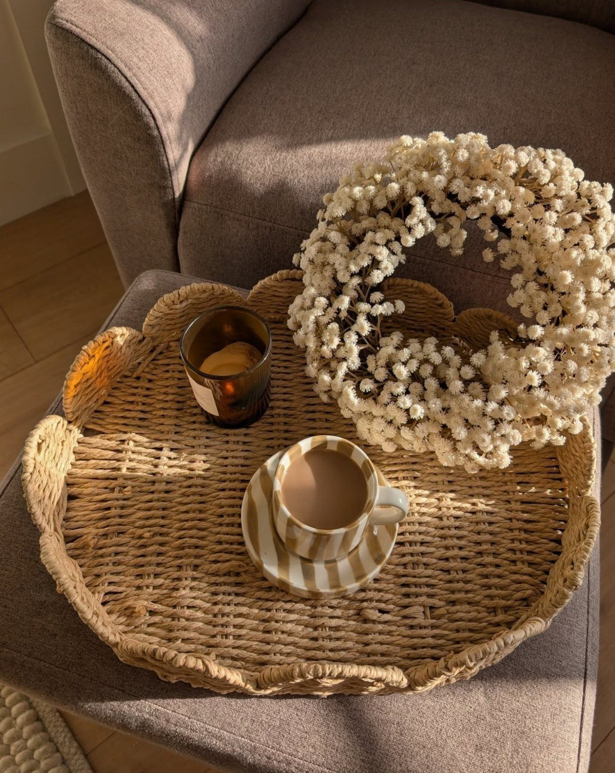 Woven wicker tray with a white flower wreath, candle, and coffee on a grey chair.