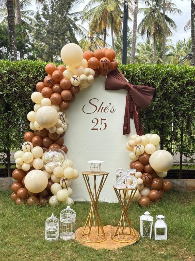 Birthday decoration with brown and cream balloons and gold pedestals in a garden.