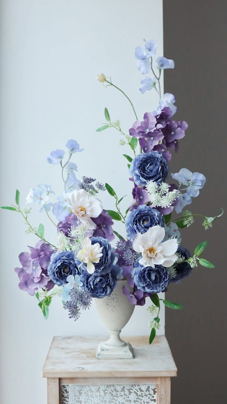 34 Large Floral Arrangement Ideas for Tables, Trays and Vases 10 Blue ranunculus and purple hydrangea arrangement in a white pedestal vase on a table.