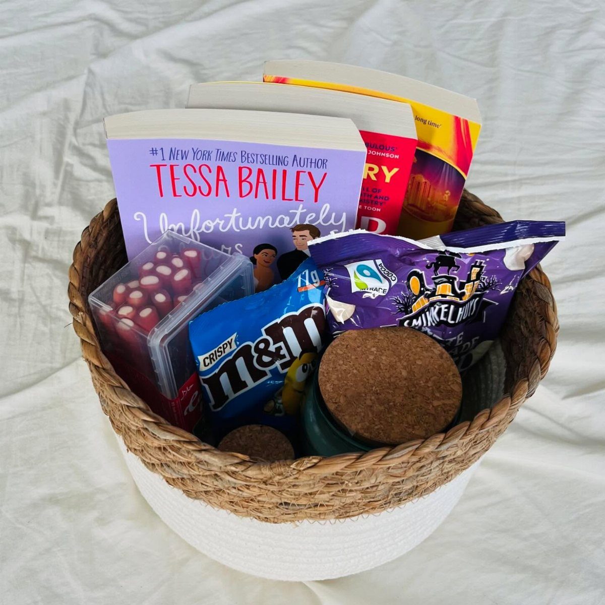 Woven gift basket filled with books, colorful candies, and cork coasters on white fabric.