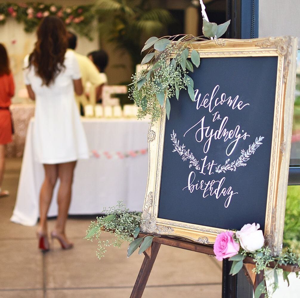 Gold framed chalkboard sign welcoming guests to Sydney's first birthday party outdoors.