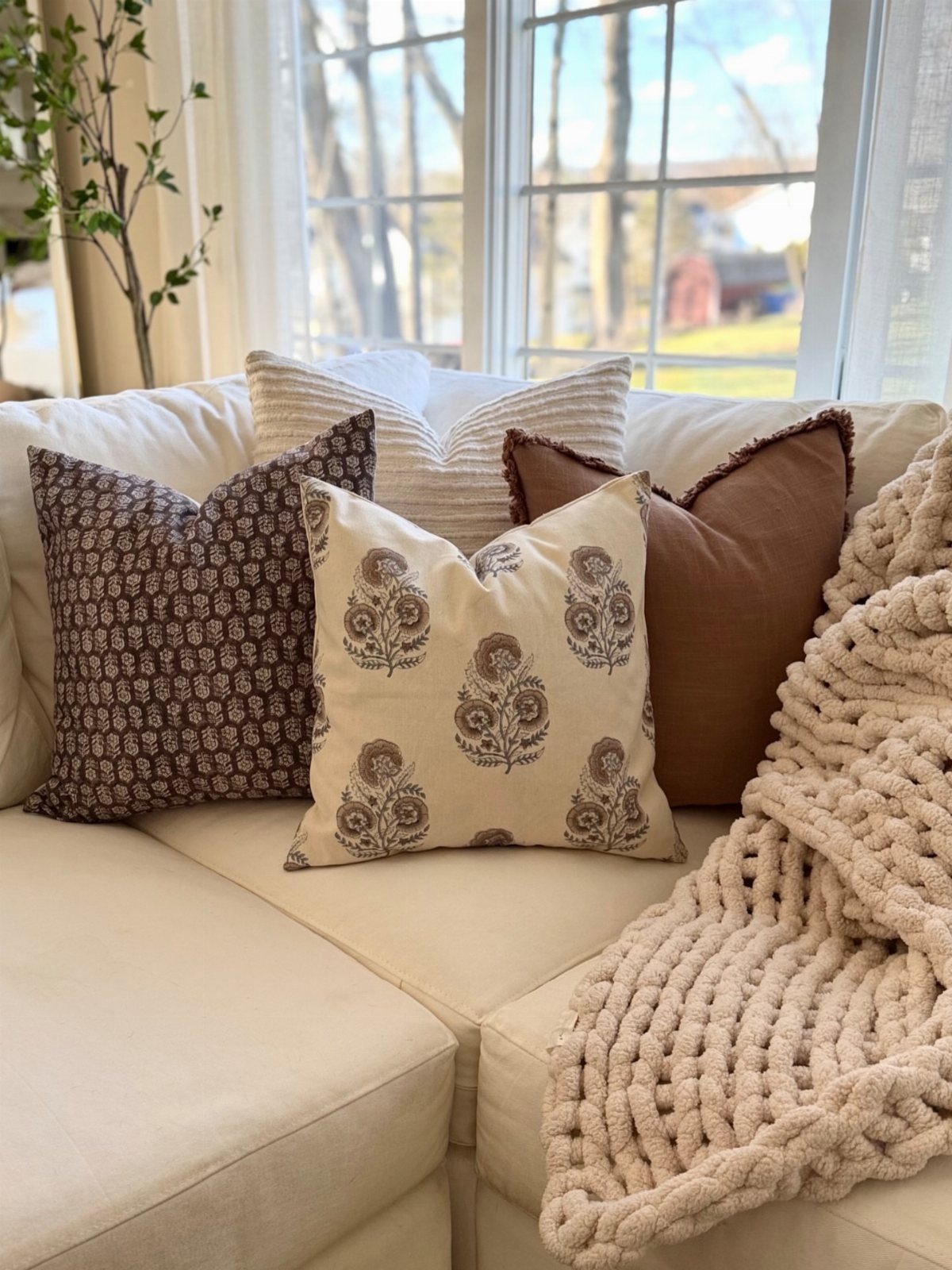 Patterned and solid brown throw pillows on a cream sofa with a chunky knit blanket.