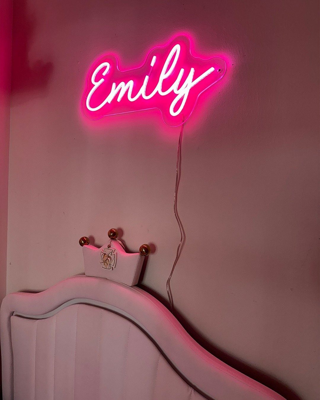 Pink neon sign spelling Emily mounted on a wall above a white bed headboard.