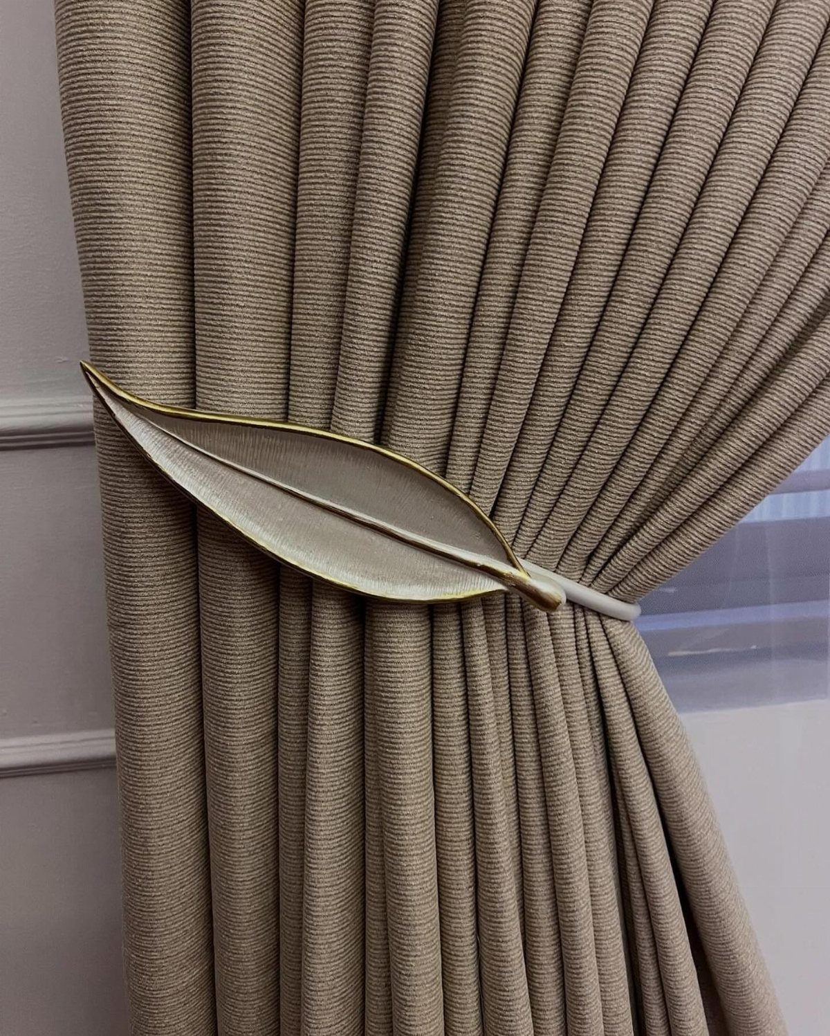 40 Home Decor Item Ideas, From Candle Holders to Sculptures 13 Gold leaf-shaped curtain tieback holding back a beige textured fabric curtain.