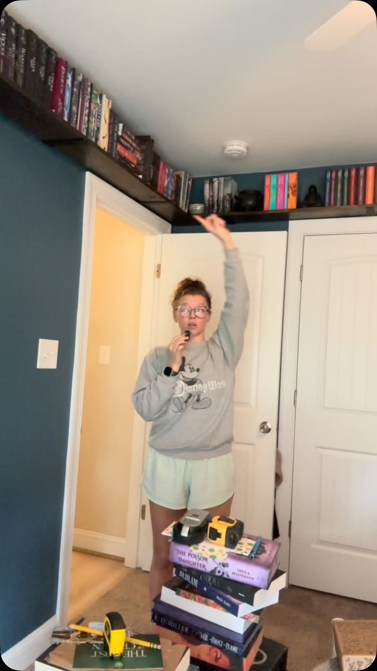 12 Ideas for Book Storage From Floating Shelves to Side Tables 7 Woman standing below a high wooden shelf filled with books framing a doorway.