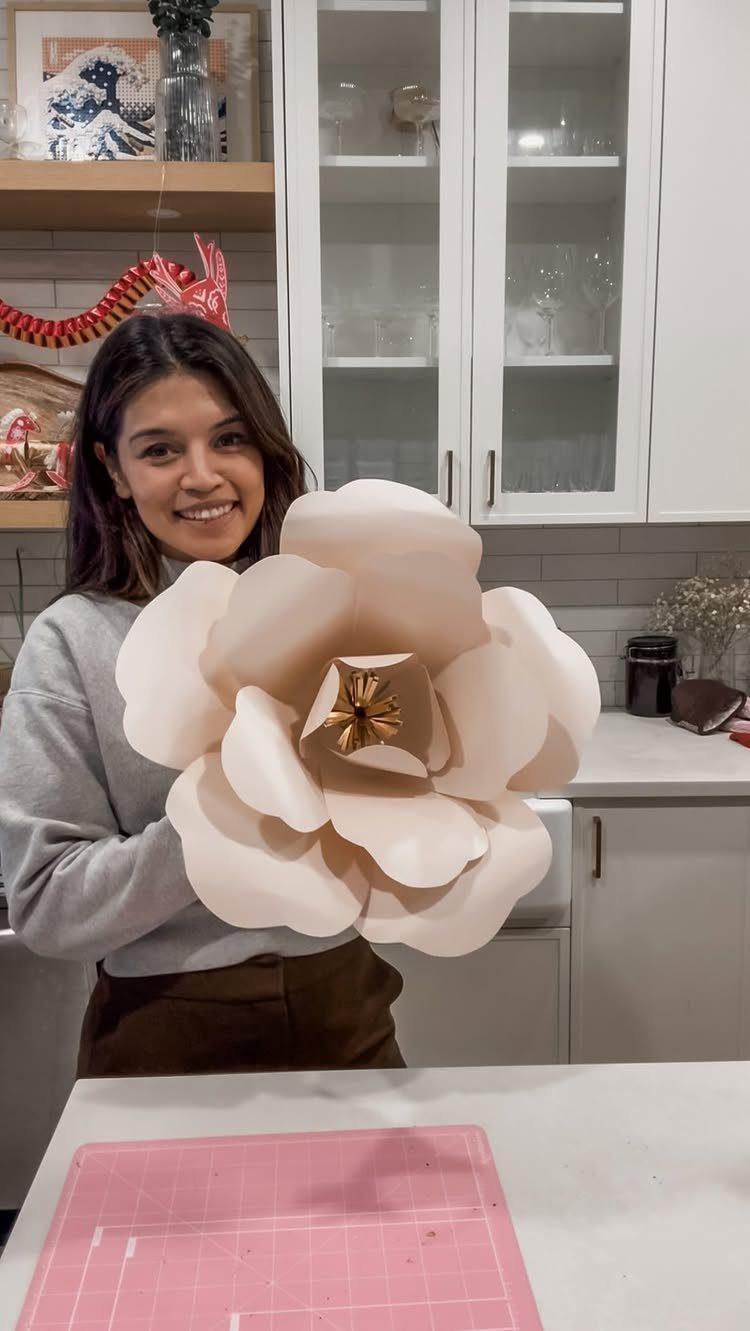 Woman holding a large pale pink paper flower in a bright kitchen.