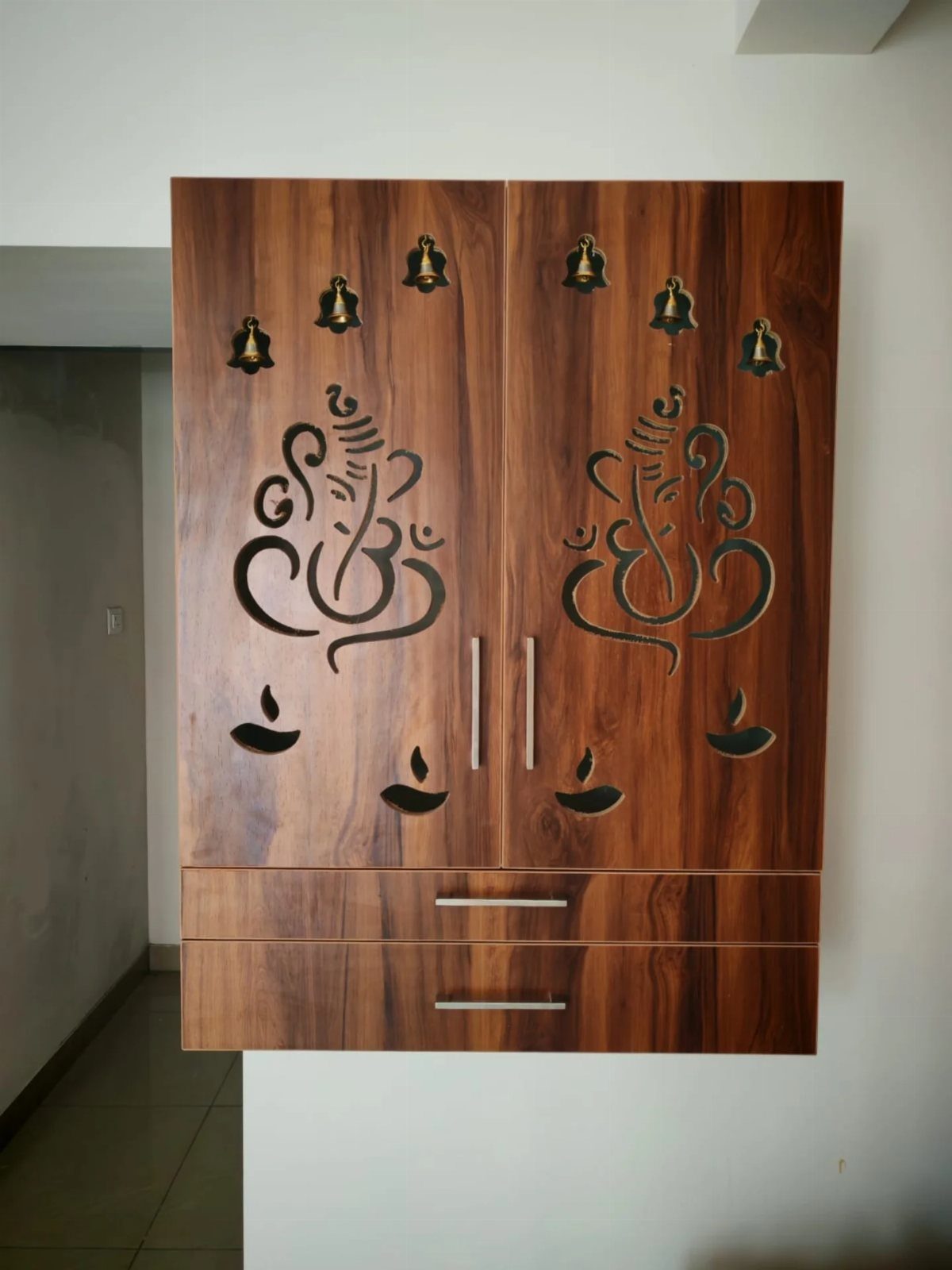34 Puja Unit Design Modern Ideas for Your Home in 2026 29 Wall-mounted wooden puja unit with carved Ganesha figures and bell cutouts on doors.
