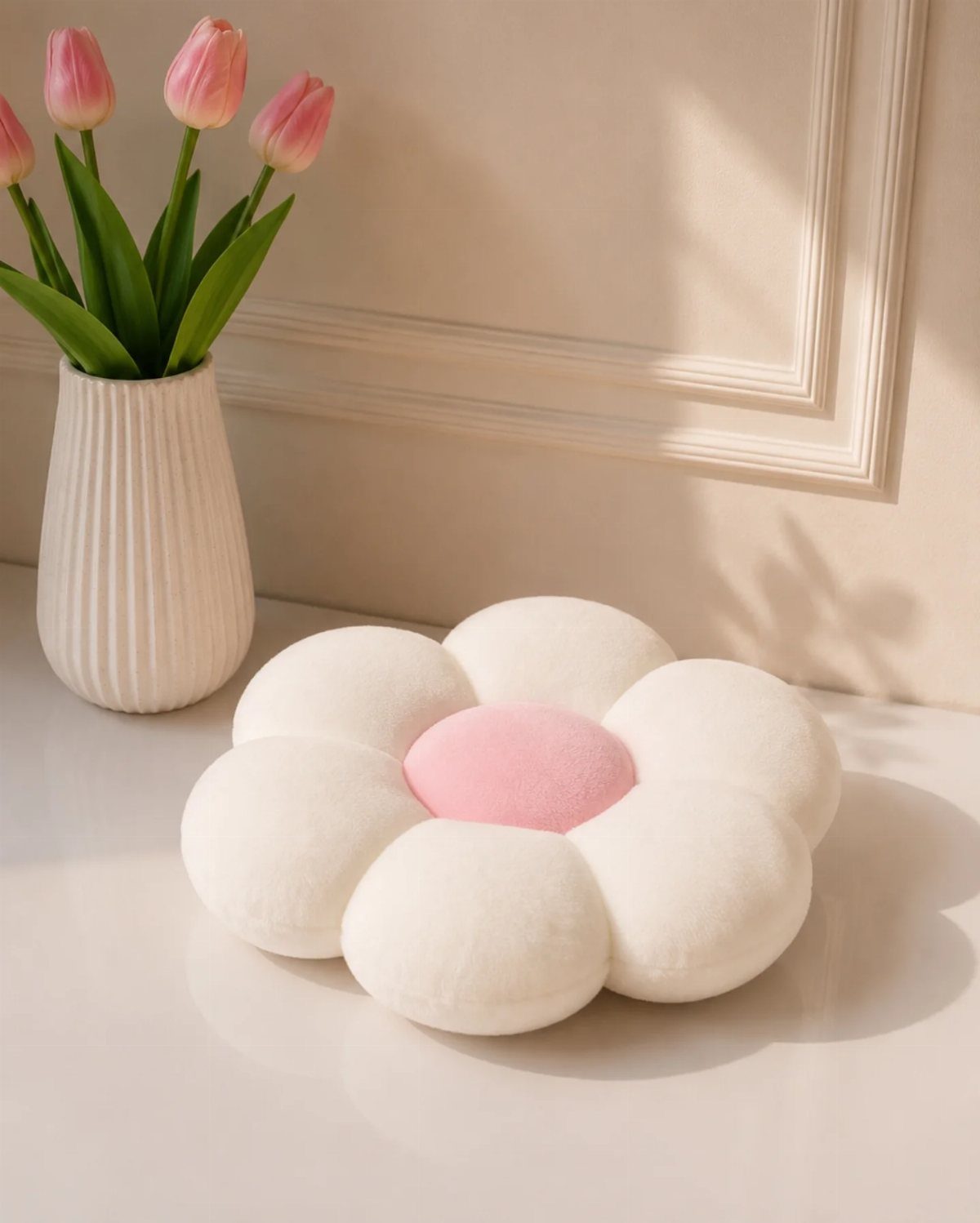 Flower-shaped white plush cushion with pink center next to pink tulips in a vase.