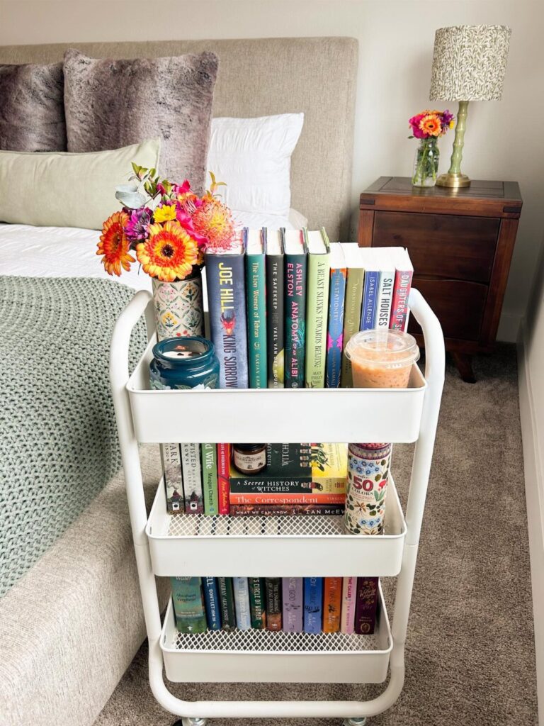 White rolling cart used as a bookshelf next to a bed in a bedroom.