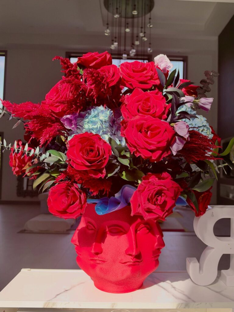 Large floral arrangement of red roses and blue hydrangeas in a red face-shaped vase.