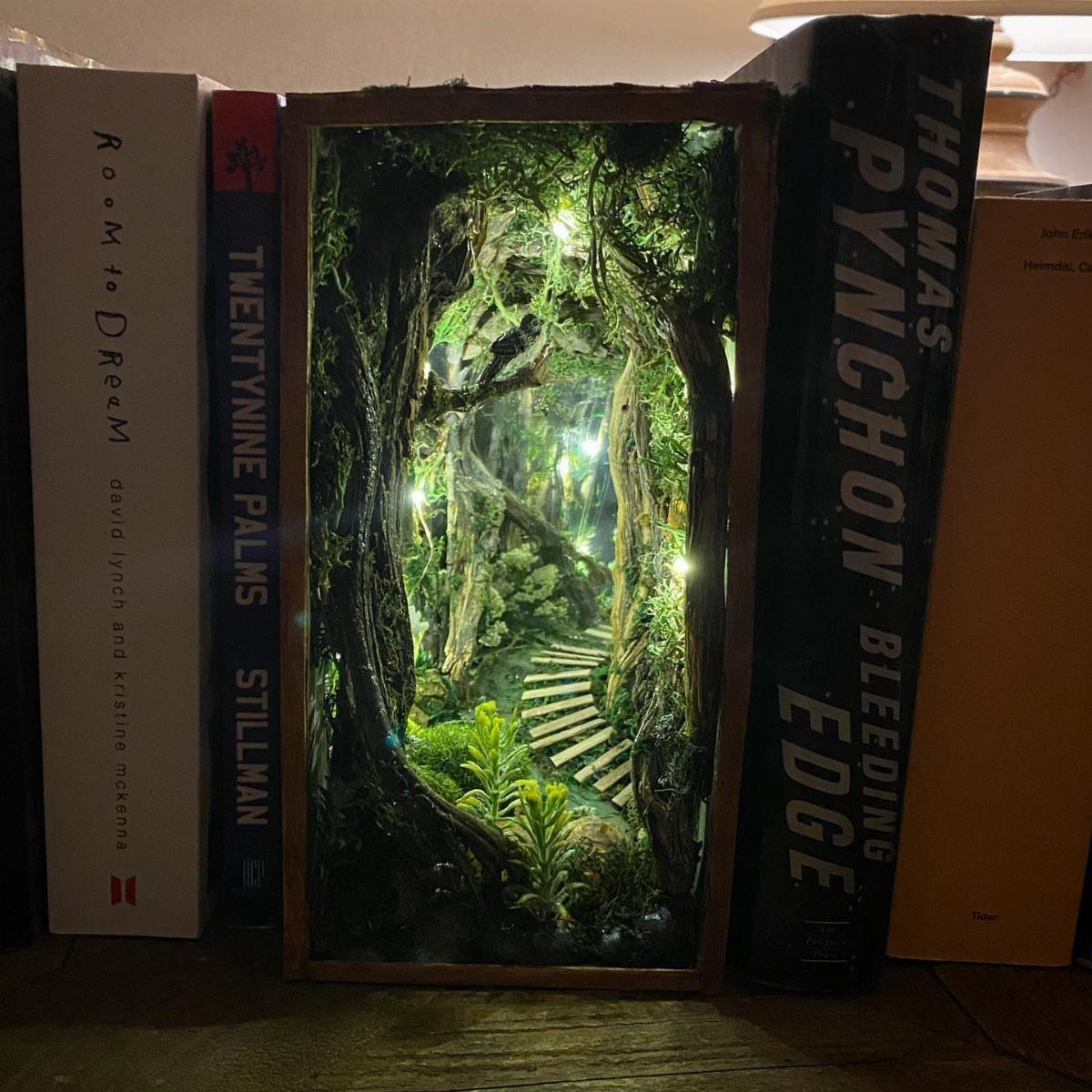 Miniature forest diorama with a wooden path and lights between books on a shelf