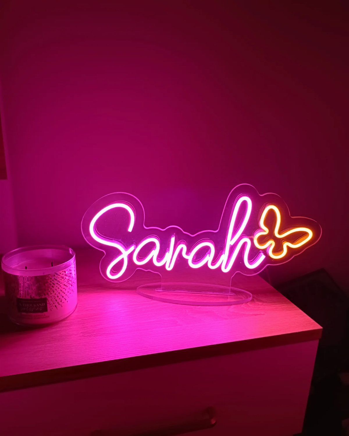 Pink neon sign spelling Sarah with a butterfly on a wooden bedside table.