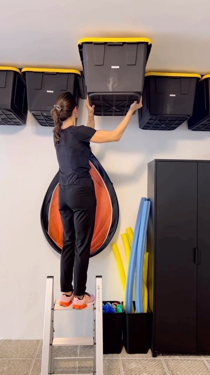 Woman on a ladder placing a black storage bin into ceiling-mounted overhead racks.