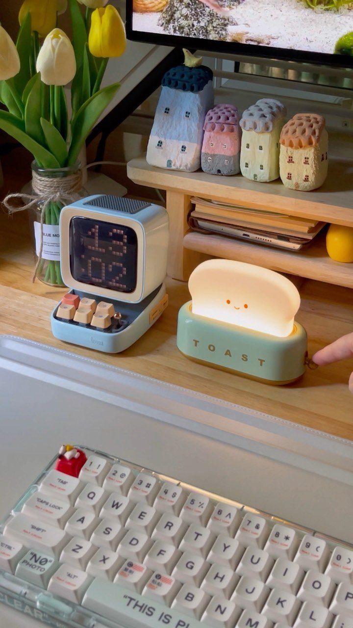 Desk setup with a toast lamp, digital clock, yellow tulips, and cute house figurines.