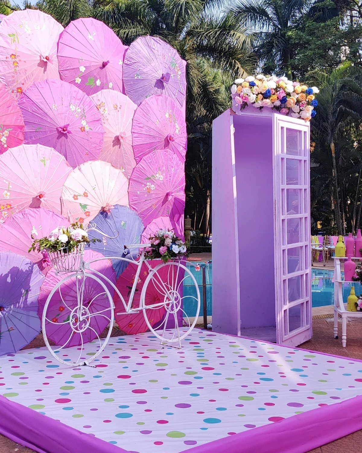 Decorative photo booth with pink umbrellas, white bicycle, and purple door outdoors.