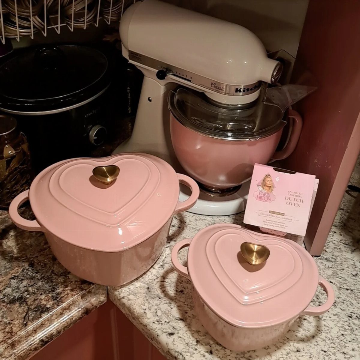 Two heart-shaped pink Dutch ovens with gold knobs on a granite kitchen counter.