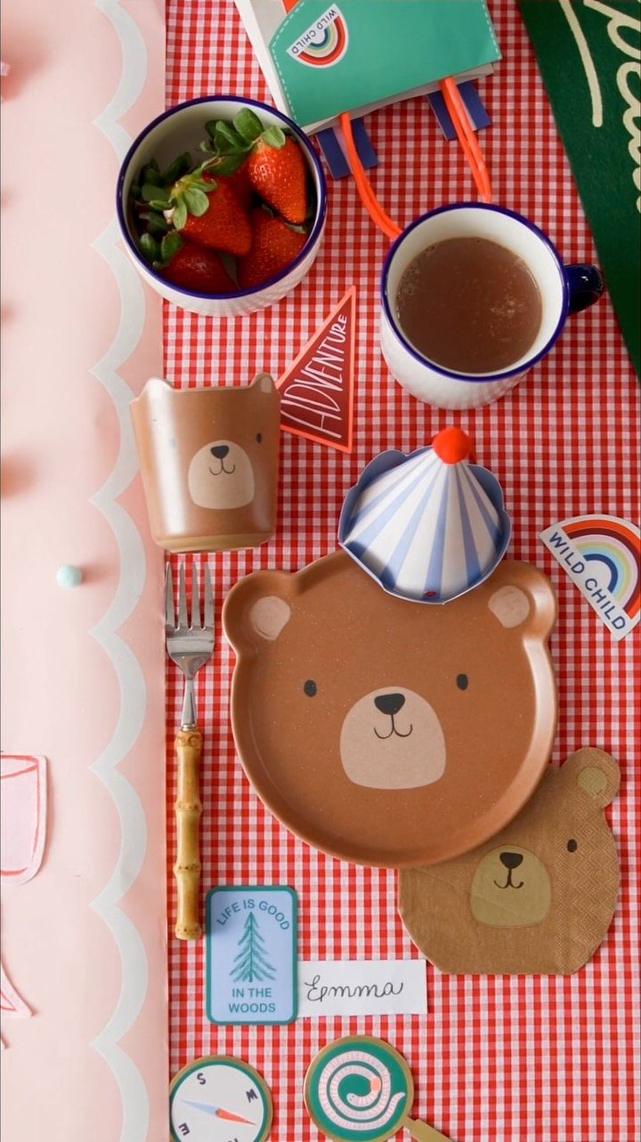 Bear-themed brown plates, cups, and cutlery on a red checkered tablecloth with party accessories.