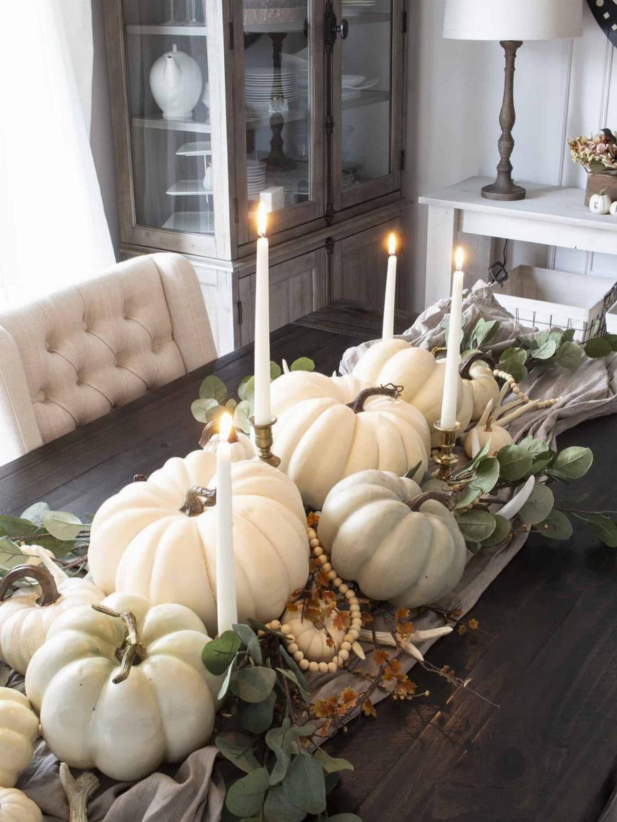 White pumpkins and tall taper candles arranged with eucalyptus on a dark wooden table.