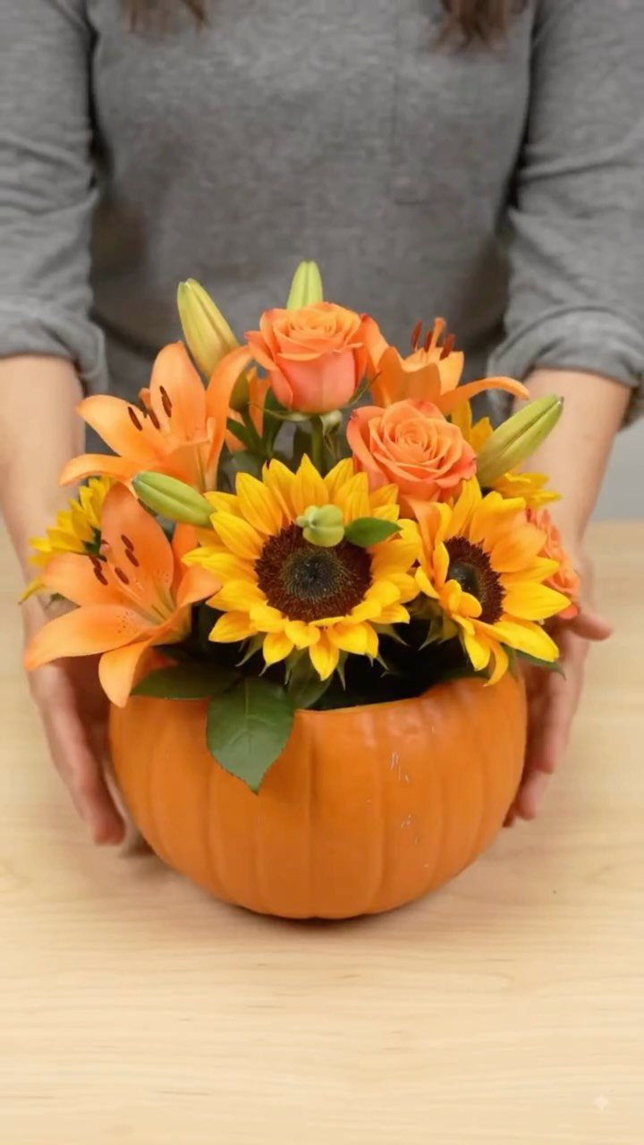 Orange pumpkin vase filled with sunflowers, orange roses, and lilies held by a person.