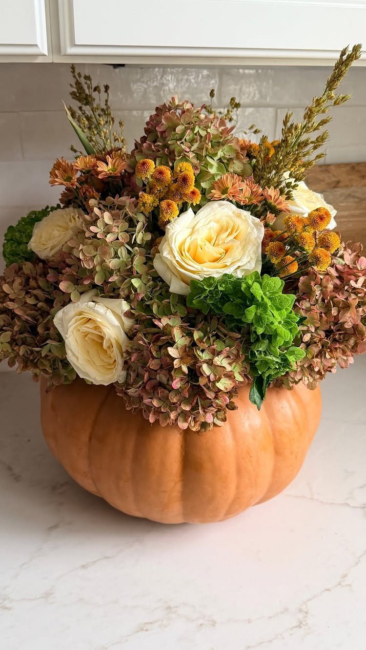 Orange pumpkin vase filled with white roses, hydrangeas, and autumn flowers on marble.