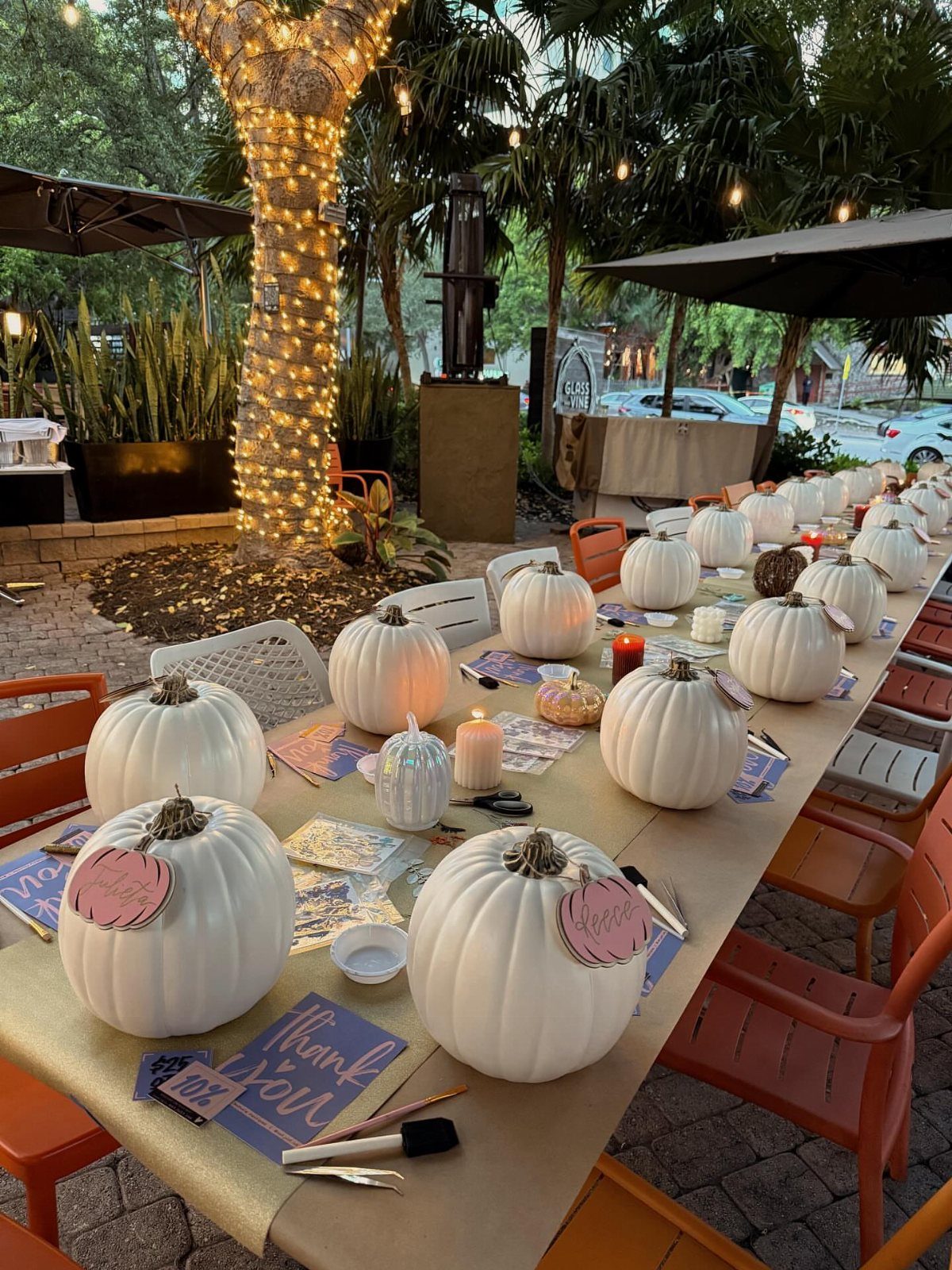 White pumpkins and candles decorating a long outdoor table with orange chairs.