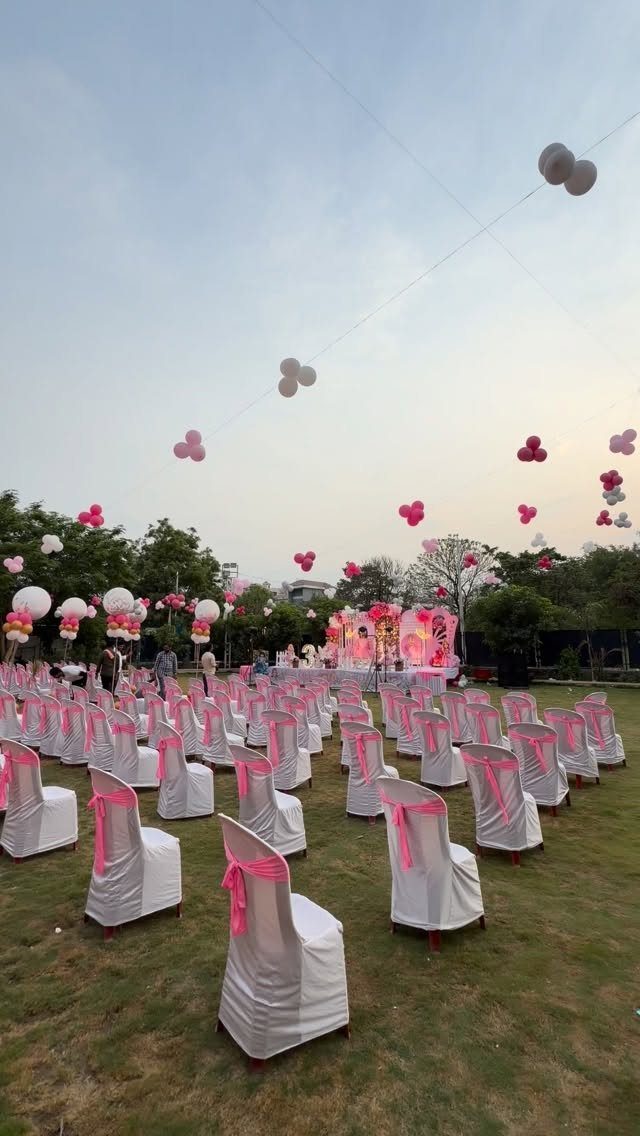 Outdoor party setup with white chairs, pink ribbons, and floating pink and white balloons.