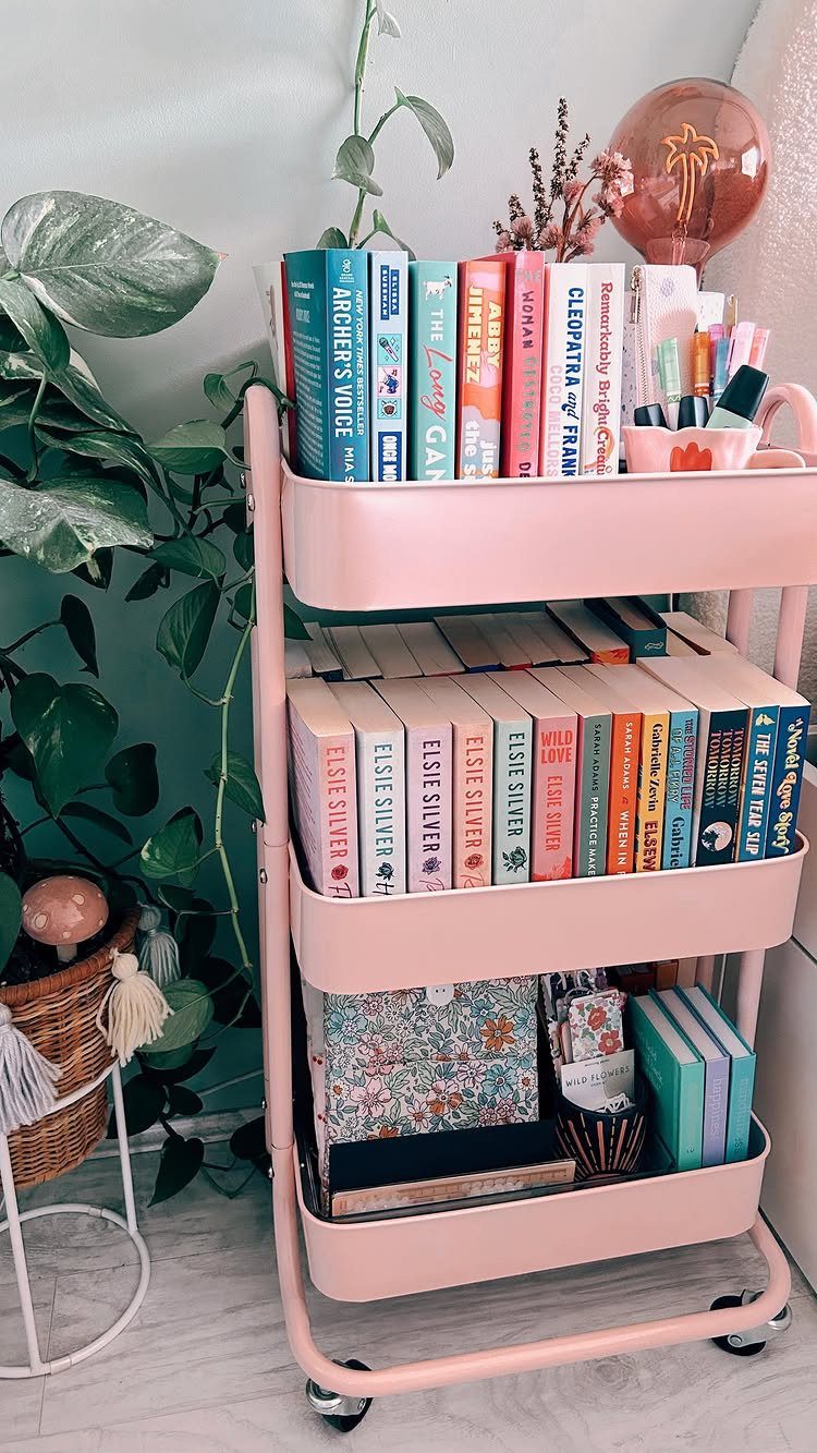 12 Ideas for Book Storage From Floating Shelves to Side Tables 2 Pink three-tier rolling cart filled with colorful books and art supplies next to a plant.