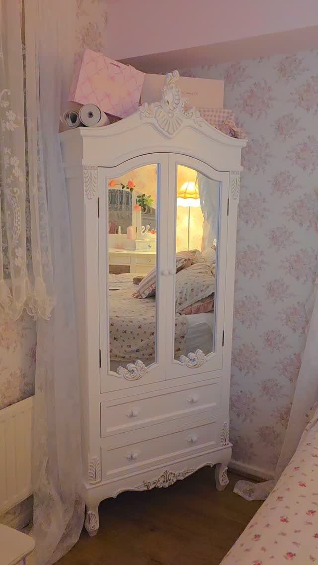 White ornate armoire with mirrored doors in a room with floral wallpaper and lace.