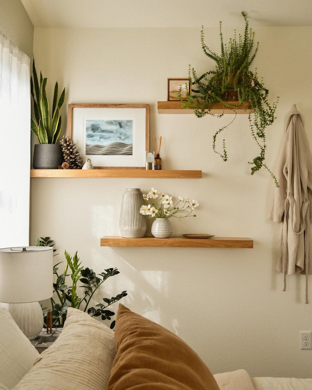 22 Bedroom Empty Wall Ideas, from Gallery Walls to Plaster 4 Three staggered wooden floating shelves on a cream wall with plants and artwork.
