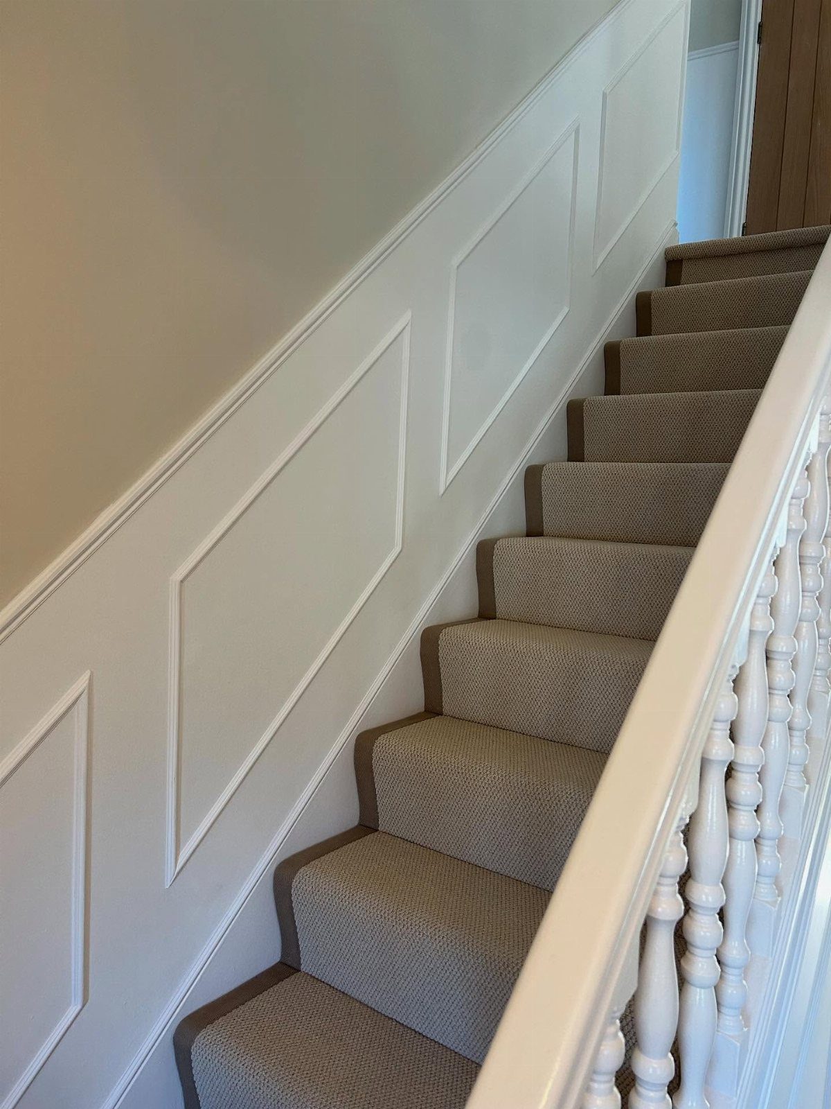 Beige carpeted stairs with white wooden banisters and white wall paneling.