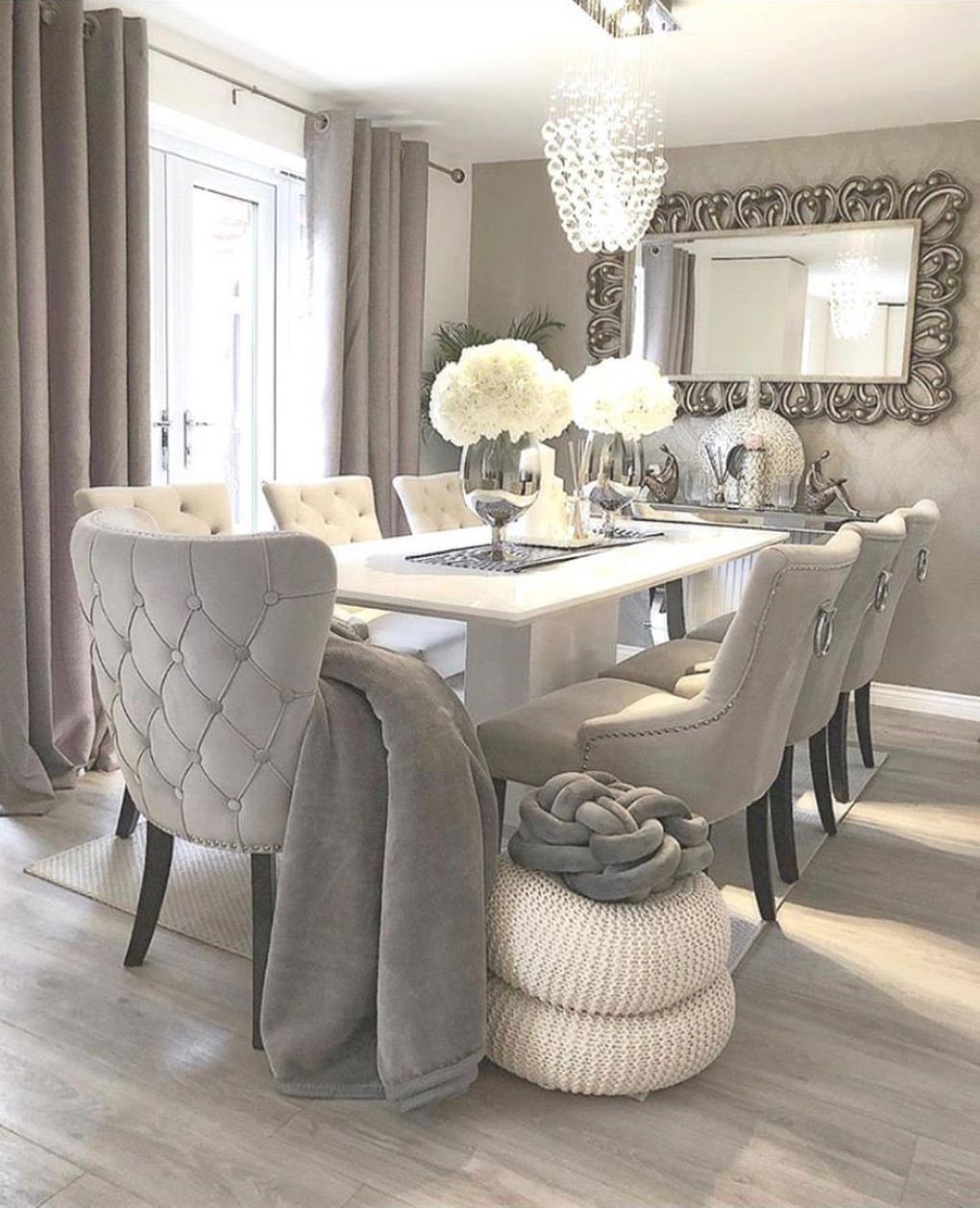 Crystal chandelier hanging over a white dining table with grey tufted chairs.