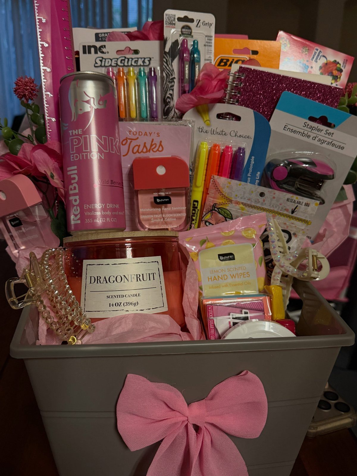 Pink themed gift basket with stationery, a candle, and energy drink in grey bin.