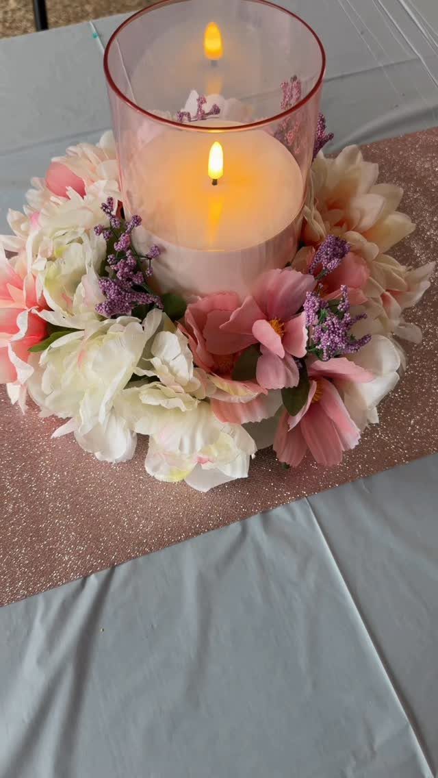 White candle in a glass cylinder surrounded by pink and white flowers on a table.