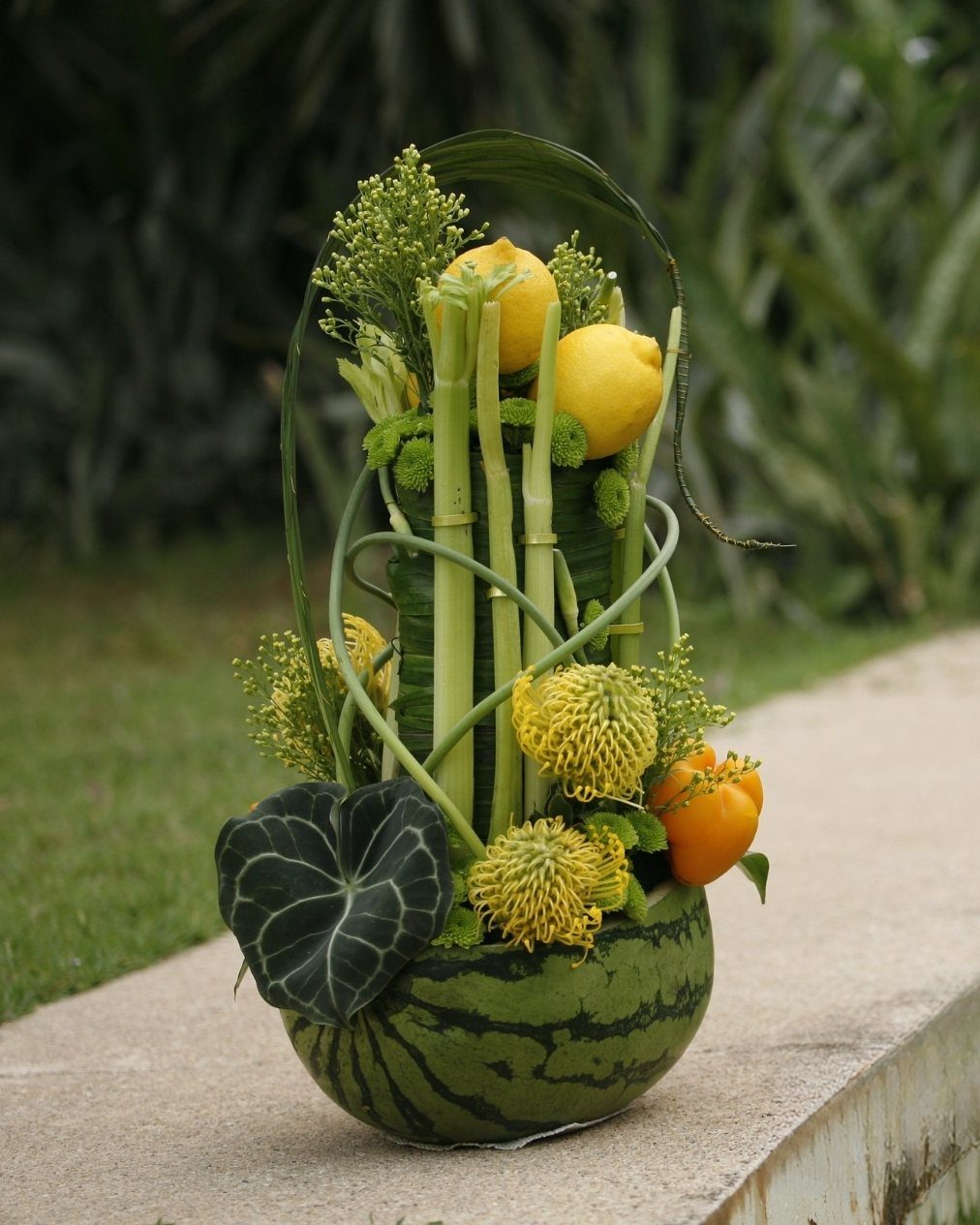34 Large Floral Arrangement Ideas for Tables, Trays and Vases 28 Floral arrangement with lemons and yellow flowers in a watermelon base outdoors.