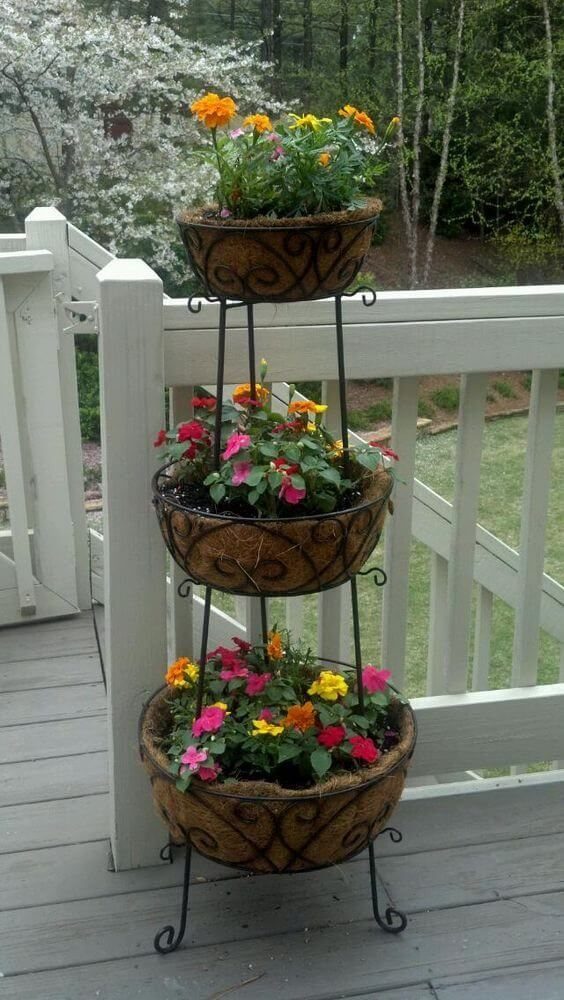 38 Flower Pot on Stand Ideas, from Iron Frames to Pedestals 8 Three-tier black metal stand with woven baskets of colorful flowers on a deck.