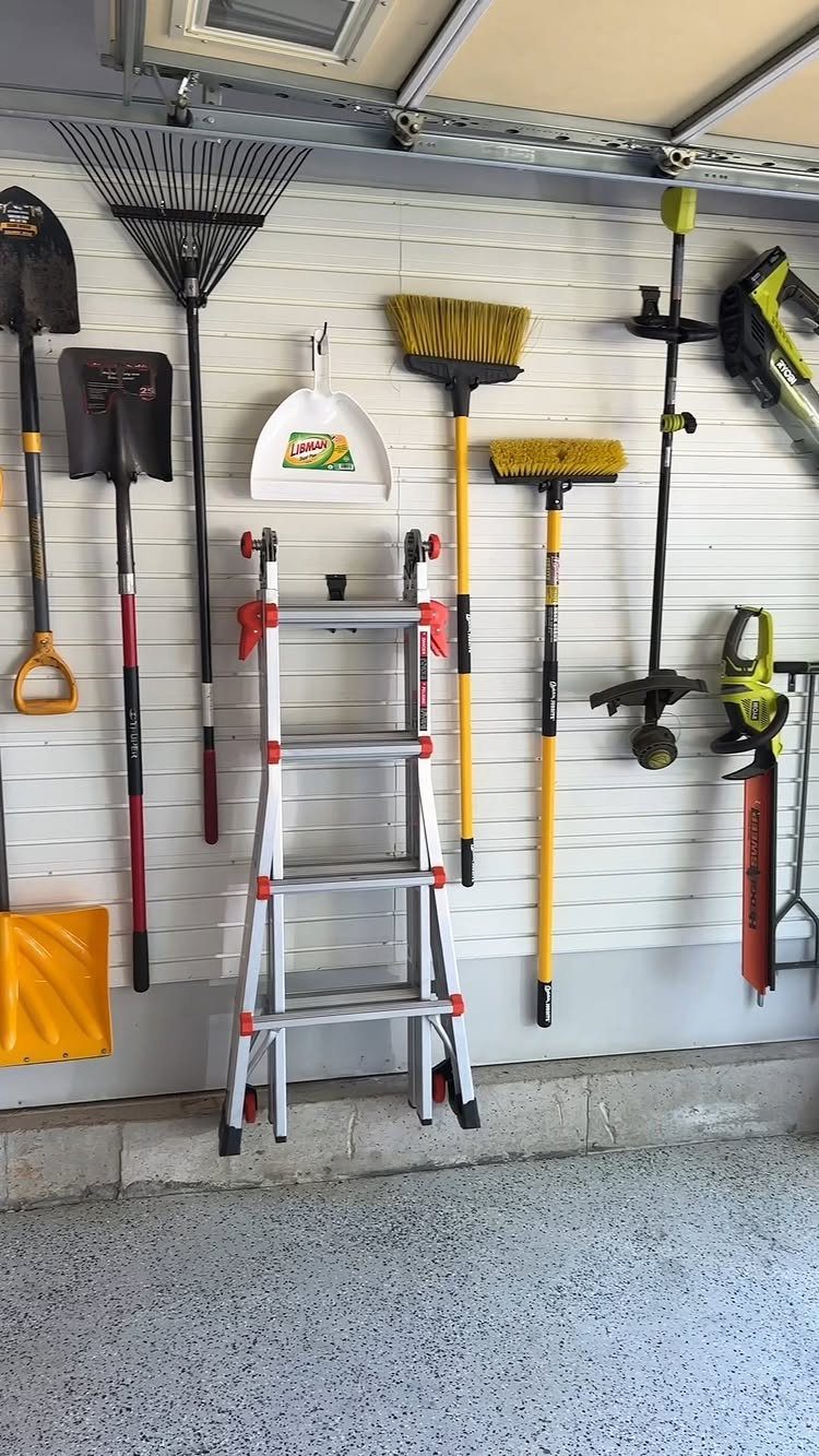 Gardening tools and a step ladder organized against a white paneled garage wall.