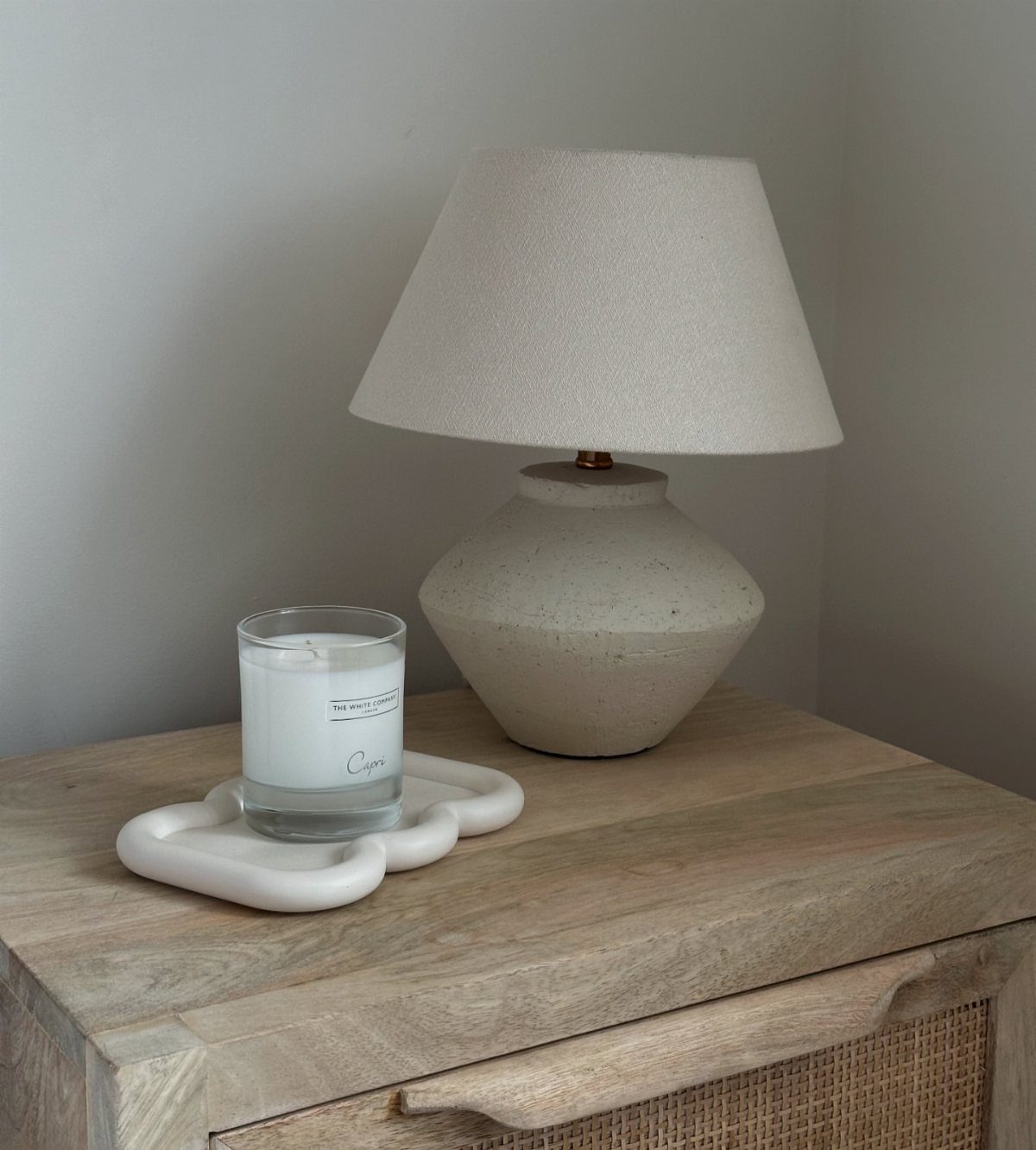 Wavy white ceramic candle tray on a light wood dresser next to a lamp.