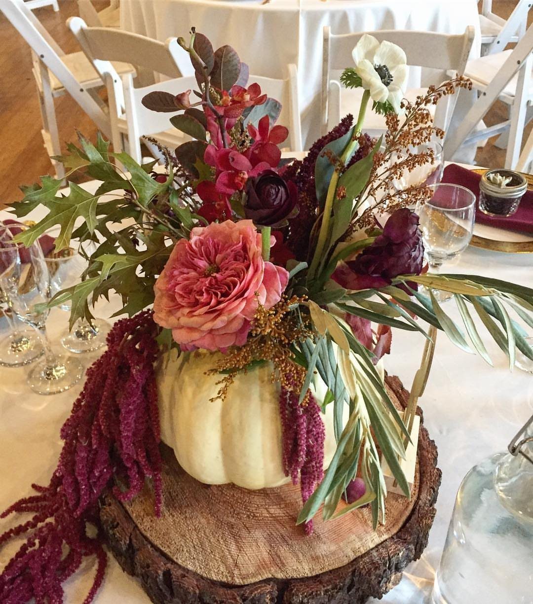 White pumpkin vase filled with pink and burgundy flowers on a wooden slice.