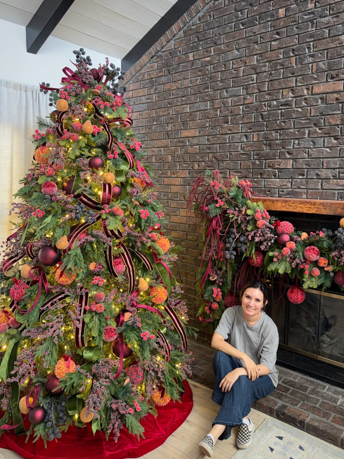 2026 Christmas Theme Ideas: 12 Looks, Heritage to Woodland 7 Woman sitting by a Christmas tree and mantel decorated with fruit and berries.