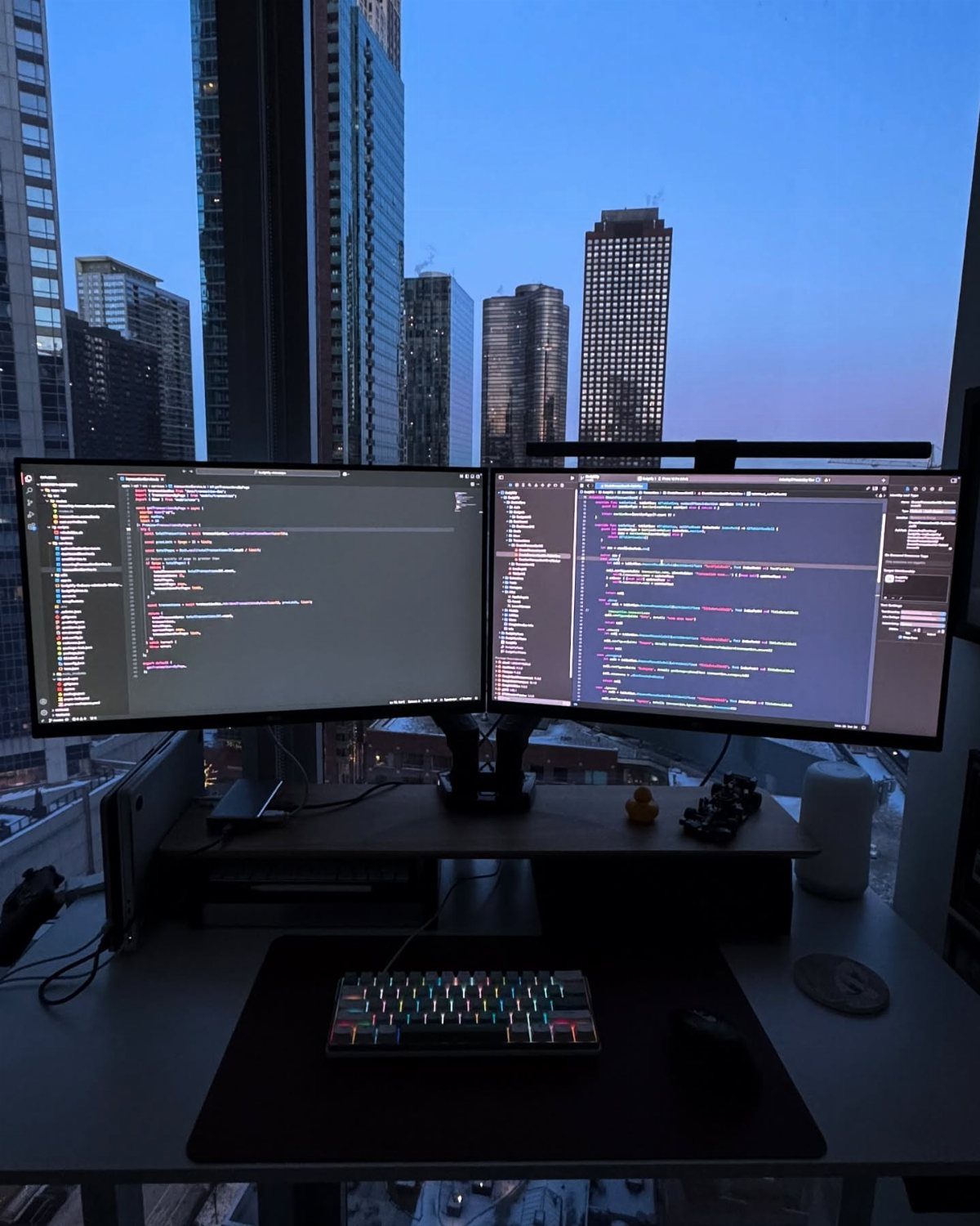 13 Double Monitor Desk Setup Aesthetic Ideas That Work 11 Double monitor desk setup with code on screens overlooking a city skyline at dusk.
