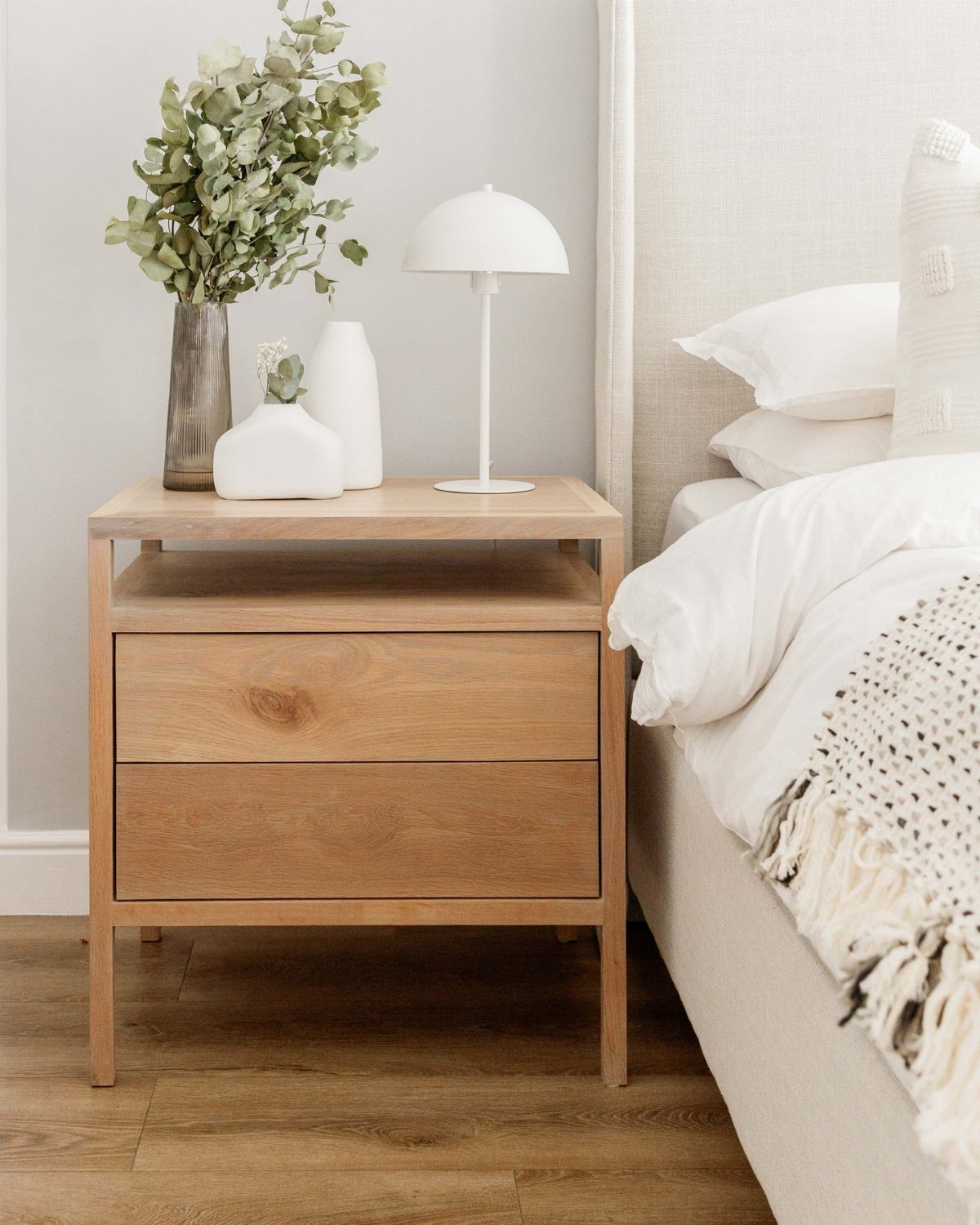 Light wood nightstand with white lamp and vases next to a white bed.