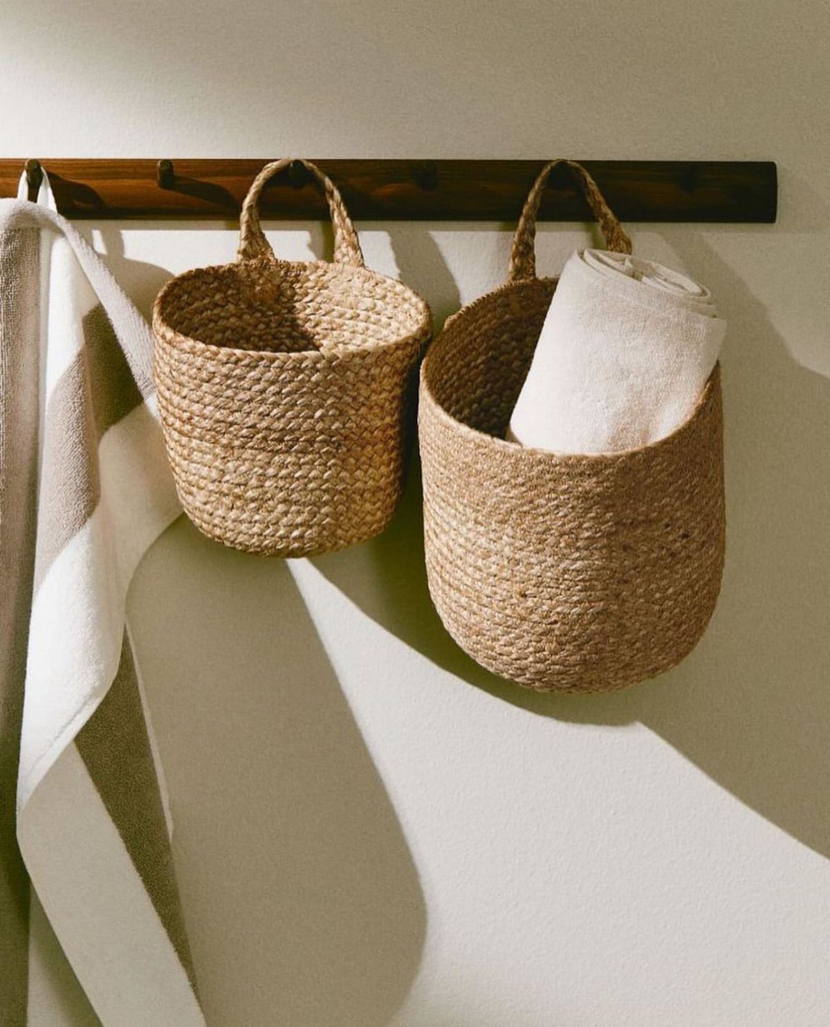 Two woven seagrass baskets hanging from a dark wood wall rack with a towel.