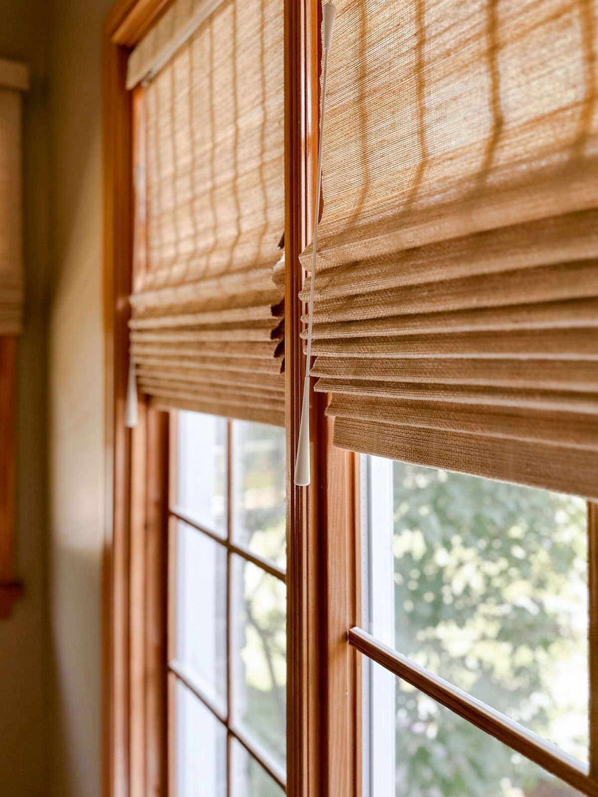 Woven wooden Roman shades partially lowered on two light brown window frames.